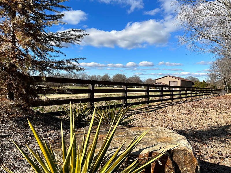 Wooden fence beside a rural field under a blue sky, with trees and a small building in the distance.