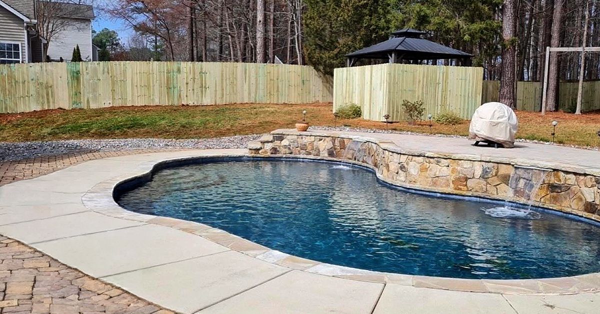 Backyard stone pool with blue water, beige patio, and wooden fence