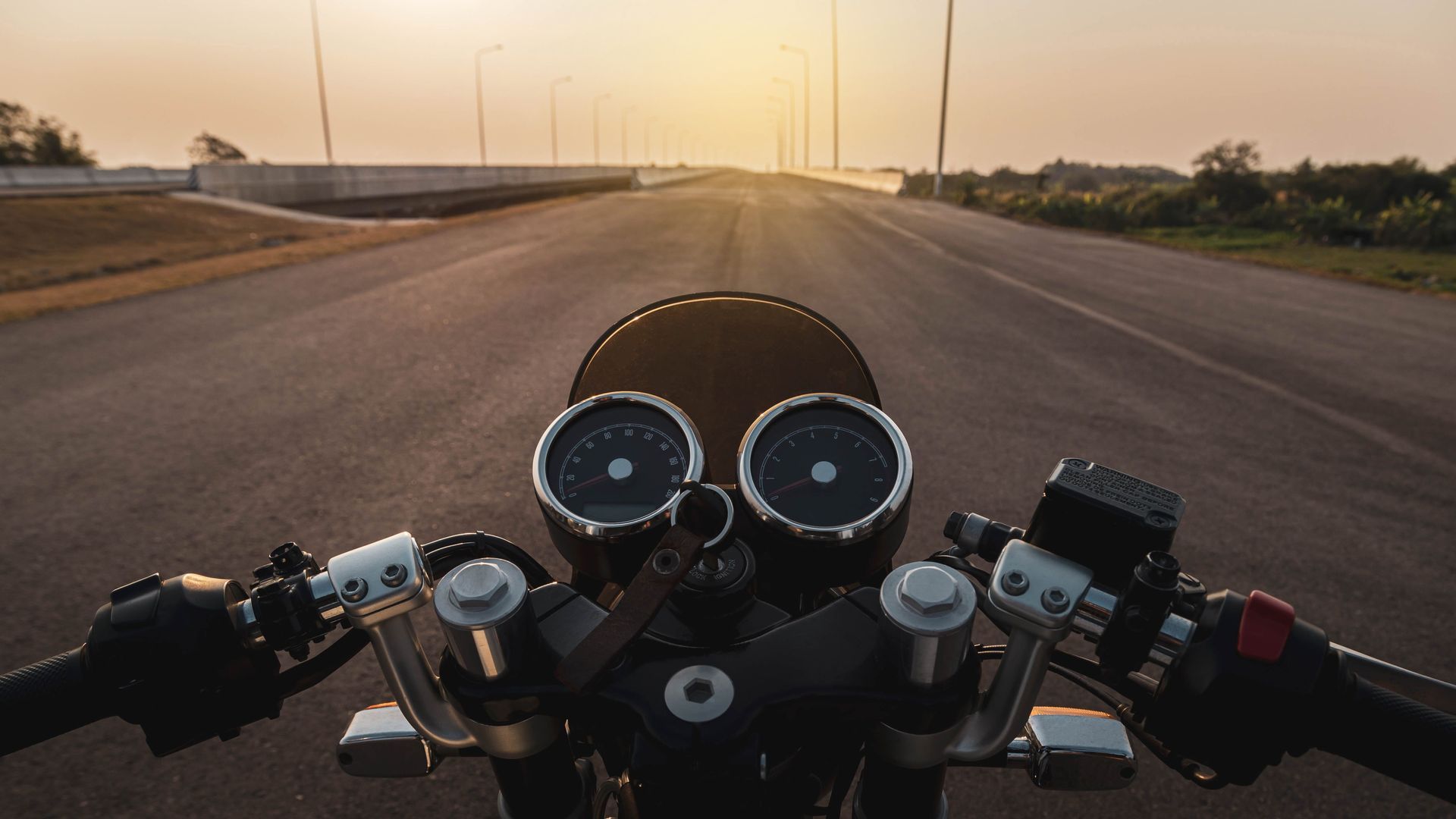 Motorcycle dashboard on an empty road at sunset, with the horizon glowing ahead.