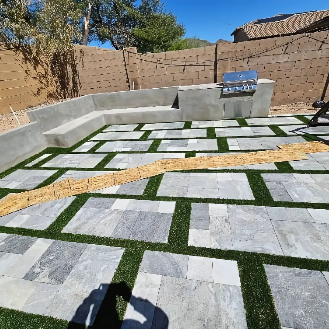 A shadow of a person is cast on a patio with grass and bricks.