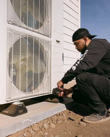 Technician kneels beside an outdoor air conditioner, inspecting wiring near a house foundation.