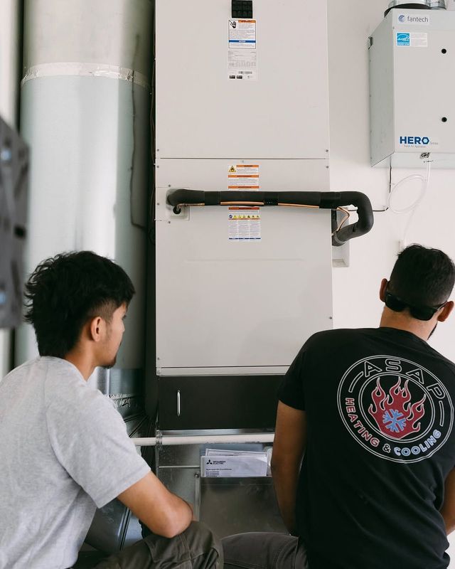 Two people standing by a wall-mounted boiler and plumbing setup indoors