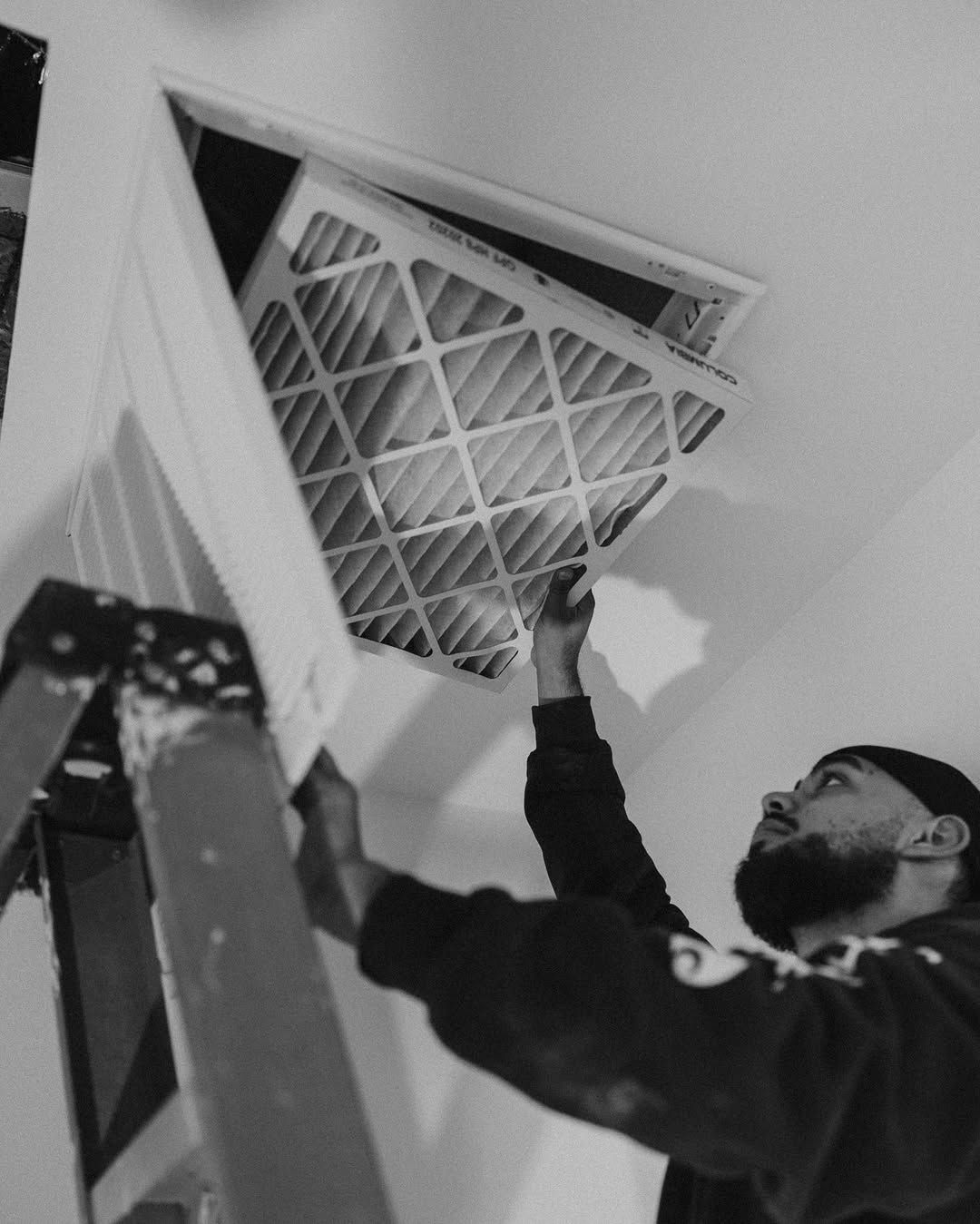 Technician replacing a ceiling air filter in a vent, reaching up from a ladder, black-and-white photo