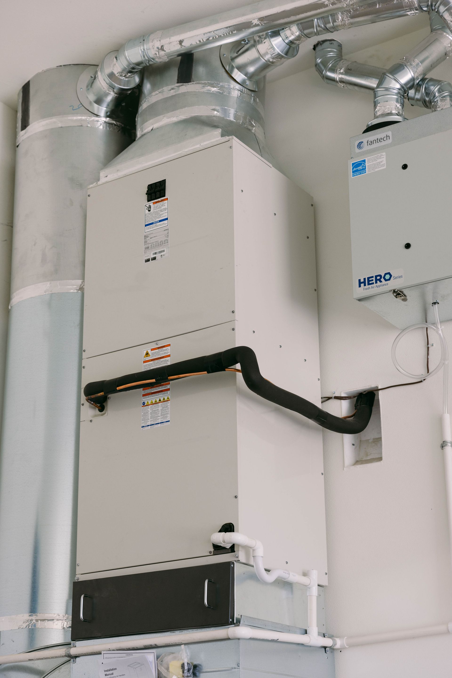 Two wall-mounted HVAC units with silver ducts and black piping in a white utility room