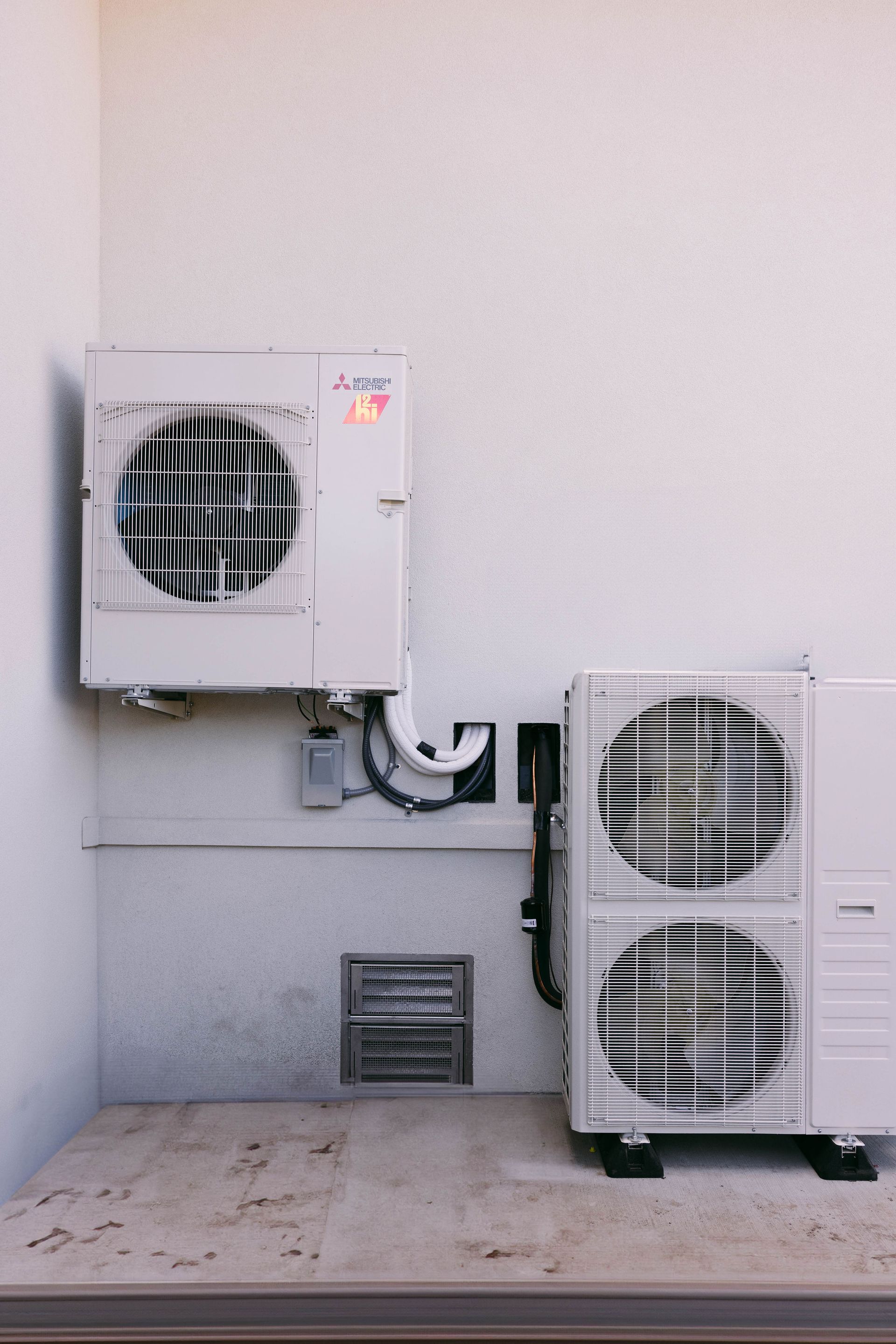 Outdoor HVAC units mounted on a wall with exposed vents and pipes on a plain exterior wall