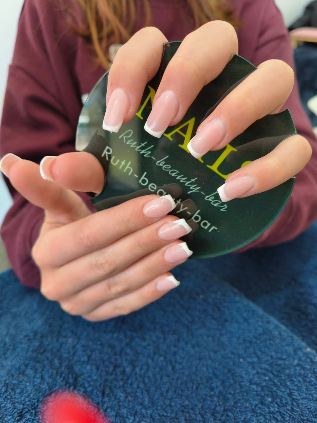 Close-up of hands displaying a fresh French manicure with clean, white tips against a circular branded nail bar card.