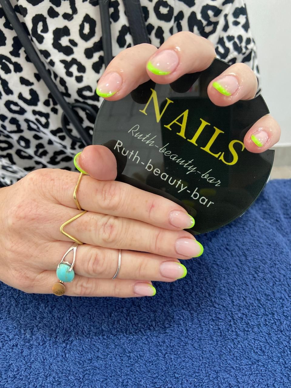 A person showing off a fresh manicure with thin, neon green French tips, holding a black promotional sign for a nail salon.