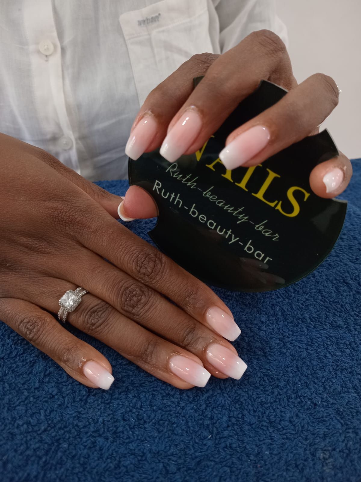 Hands displaying a pink and white ombre manicure, holding a circular black sign with gold logo text on a blue surface.