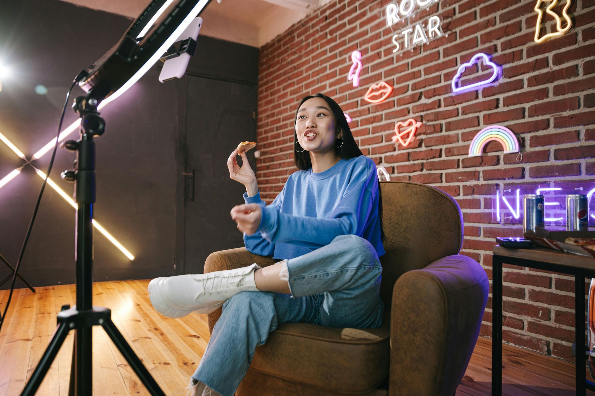 A content creator sitting in a chair, speaking while holding a snack, with neon signs on a brick wall and studio lighting.