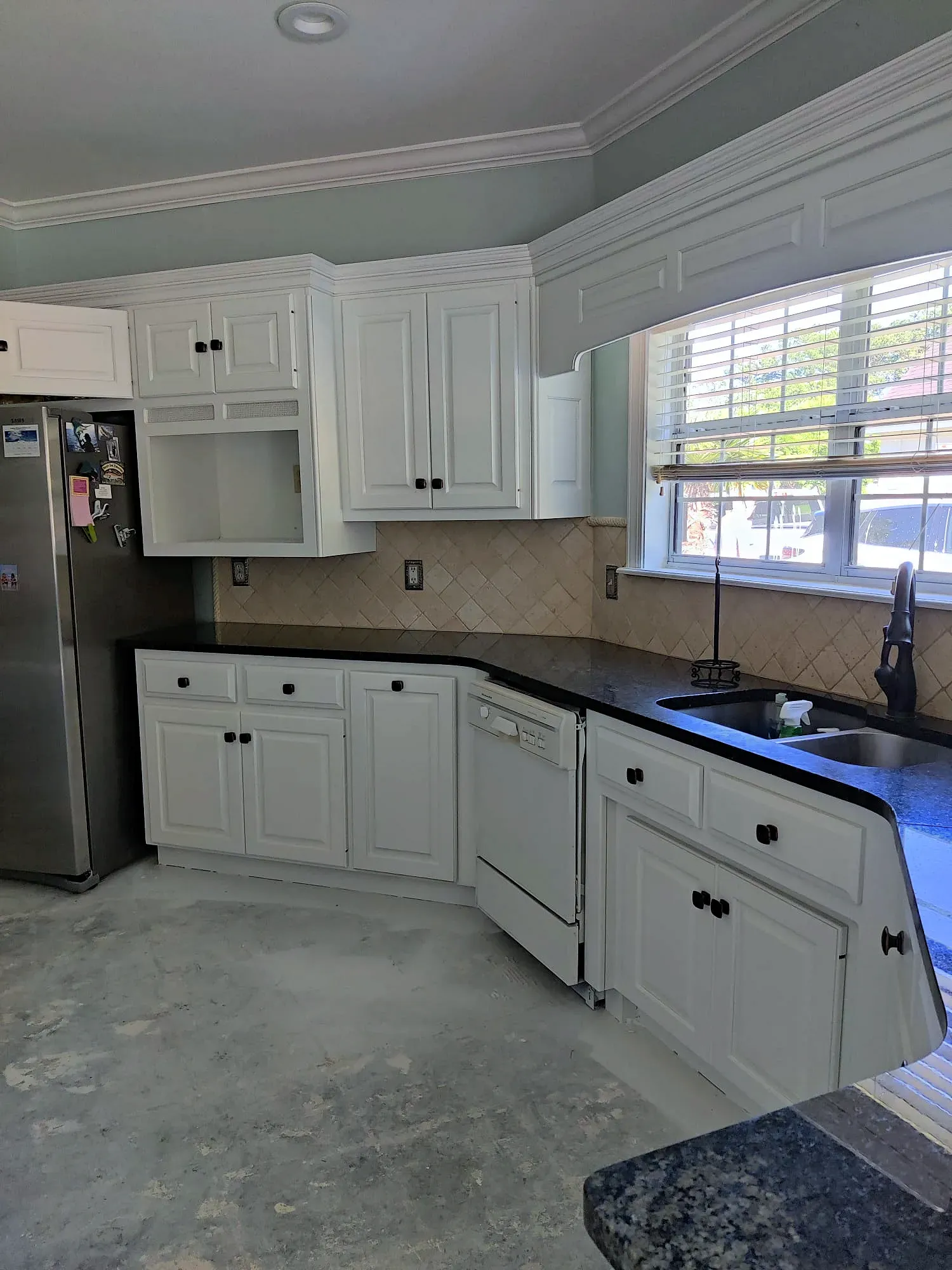 White kitchen cabinets and black countertops. Stainless steel refrigerator and dishwasher. Beige backsplash, gray floor.