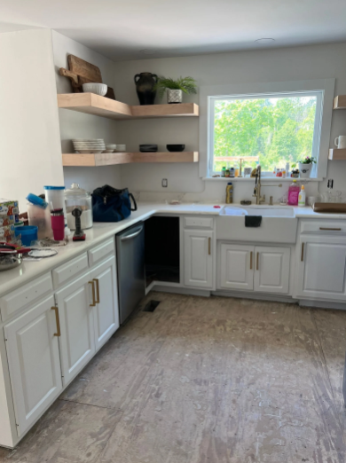 White kitchen with cabinets, dishwasher, farmhouse sink, and wooden shelves.