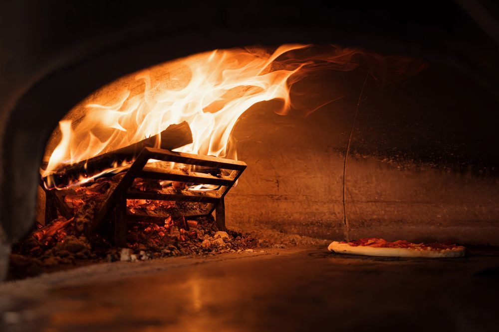 Una pizza cuoce sul pavimento di pietra di un forno a mattoni, accanto a una rastrelliera di legna ardente dalle fiamme arancioni brillanti.