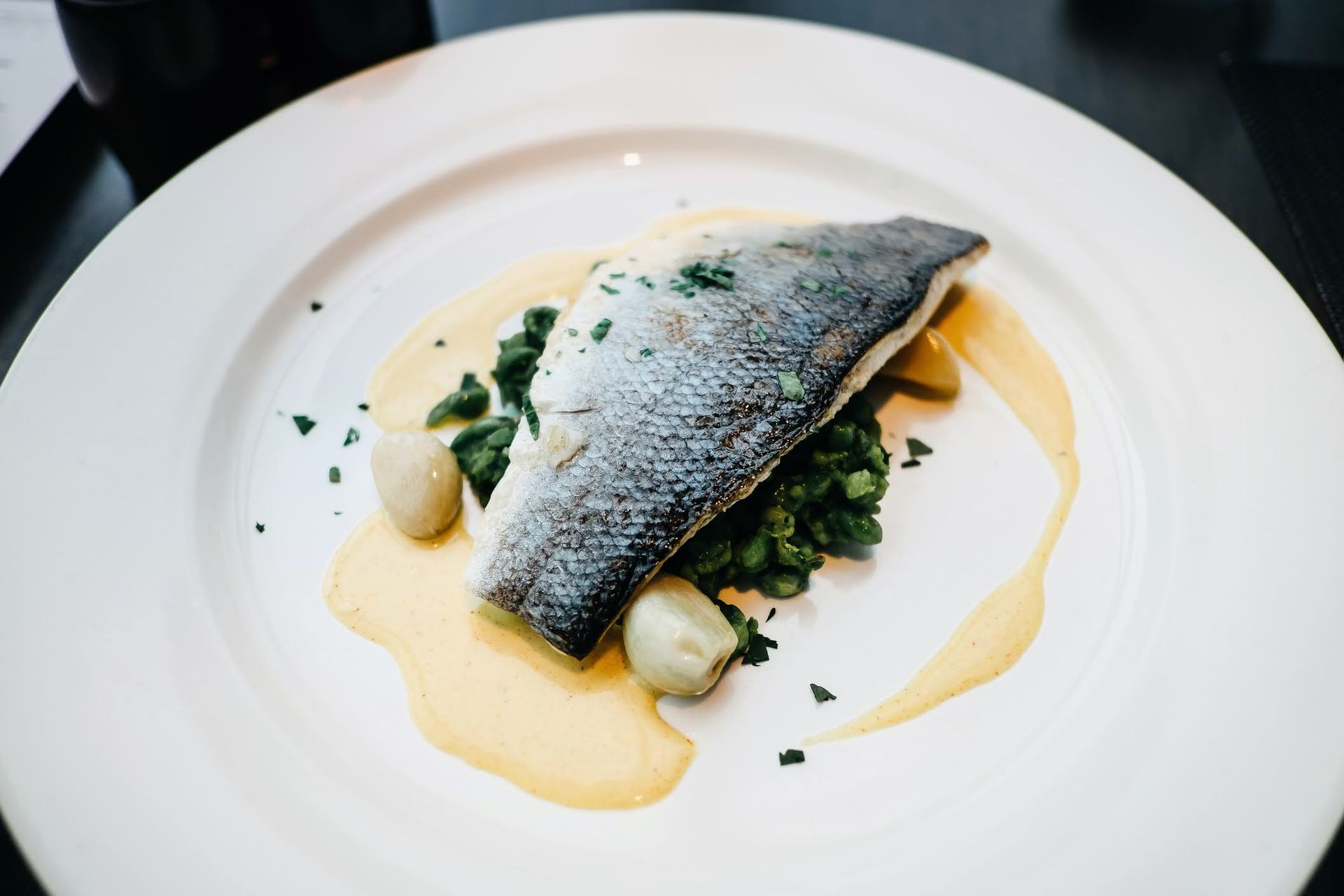 Un pezzo di pesce bianco scottato in padella con la pelle rivolta verso l'alto, servito su un letto di verdure con cipolline e una salsa gialla leggera.