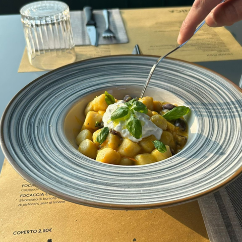 Primo piano di una mano che usa una forchetta per mangiare gnocchi conditi con basilico fresco e panna, serviti in una ciotola decorata.