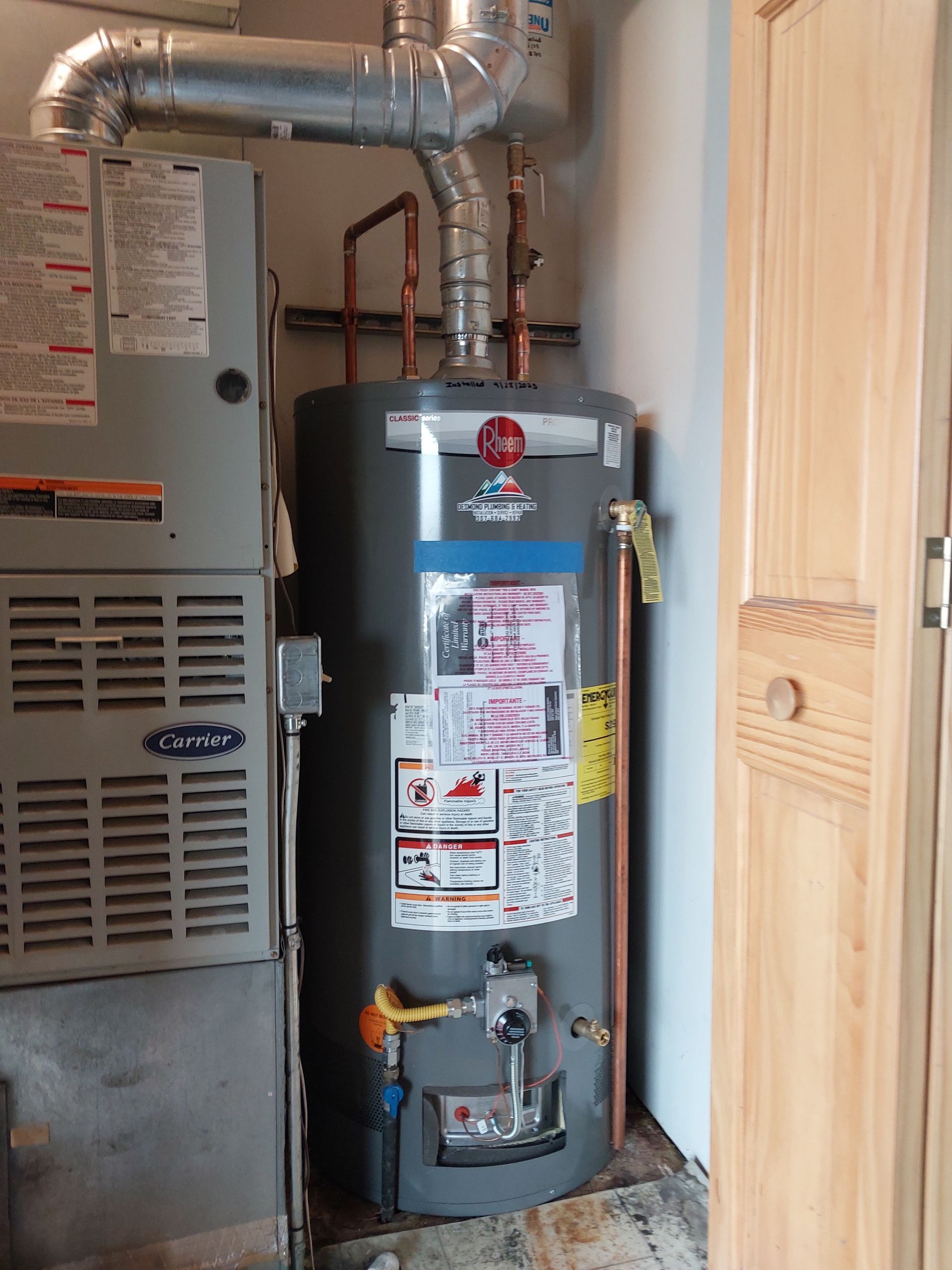 Water heater and furnace in a utility room; gray appliances, silver vent pipes, and wooden door.