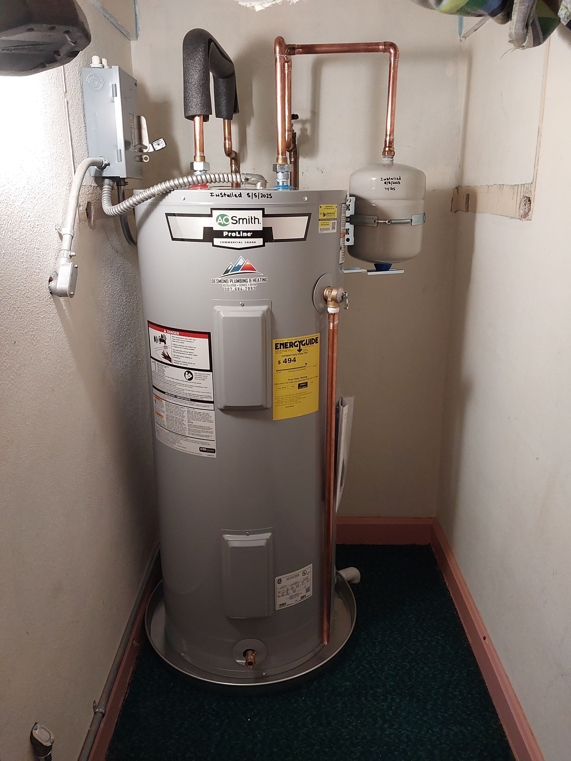 A tall, gray water heater with copper pipes in a small, enclosed space, against white walls.