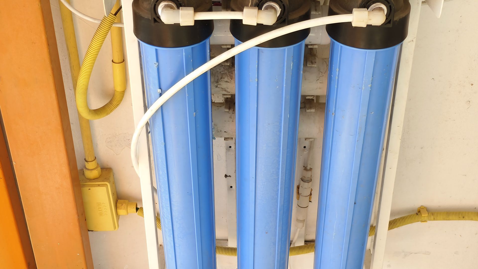 Three blue water filter housings mounted on a white surface, with white tubing and electrical wiring.