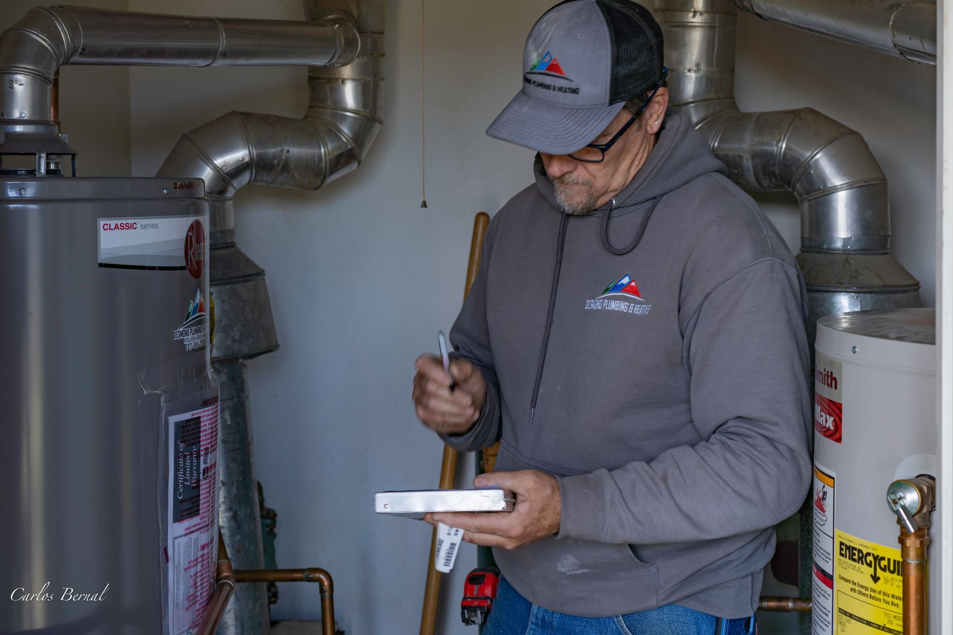 Man in grey sweatshirt inspecting water heater in utility room.