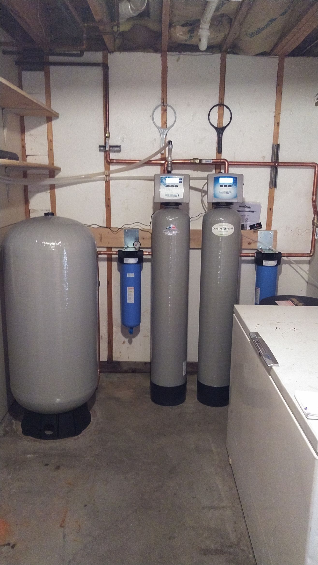 Water filtration and pressure tank system in a utility room with exposed framing and a freezer.