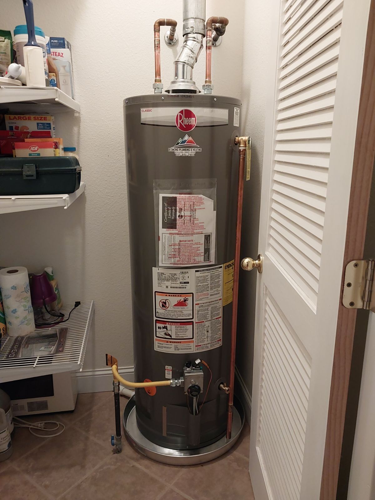 A tall, gray Rheem water heater in a utility closet with a white louvered door and shelves with supplies.