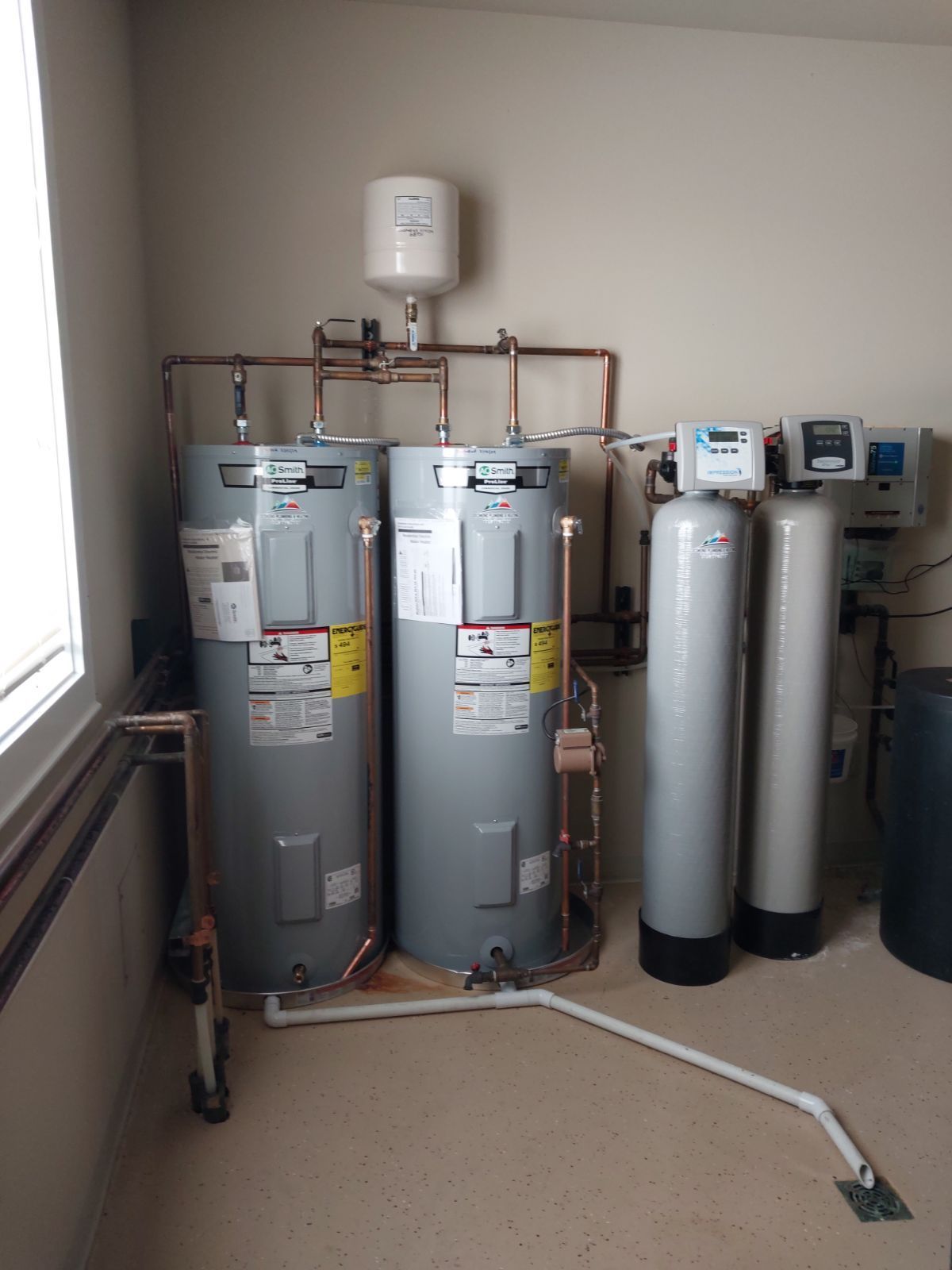 Water heater system with two tanks, copper pipes, water filters, and a white pressure tank in a room.