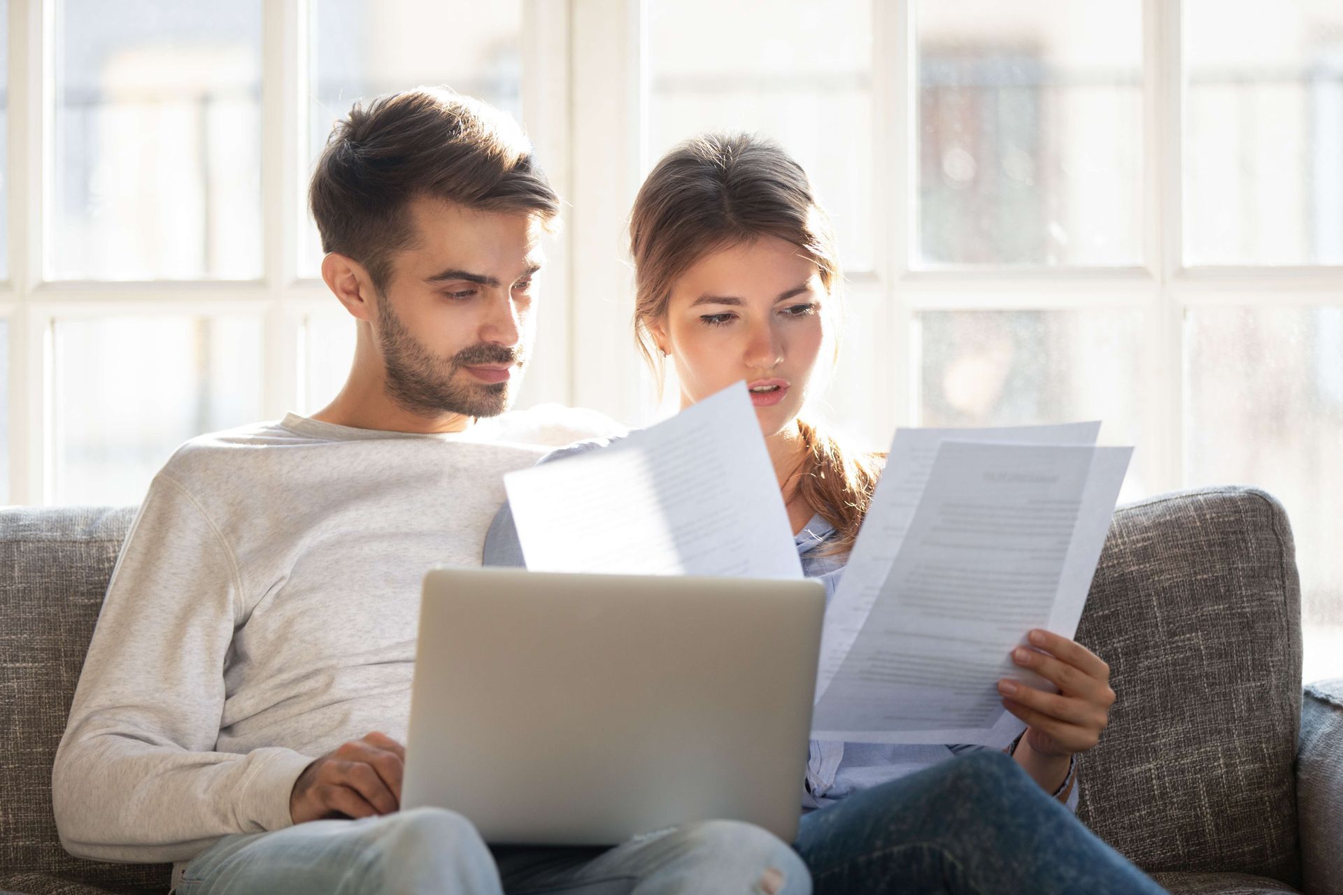 a man and a woman are sitting on a couch looking at papers and a laptop