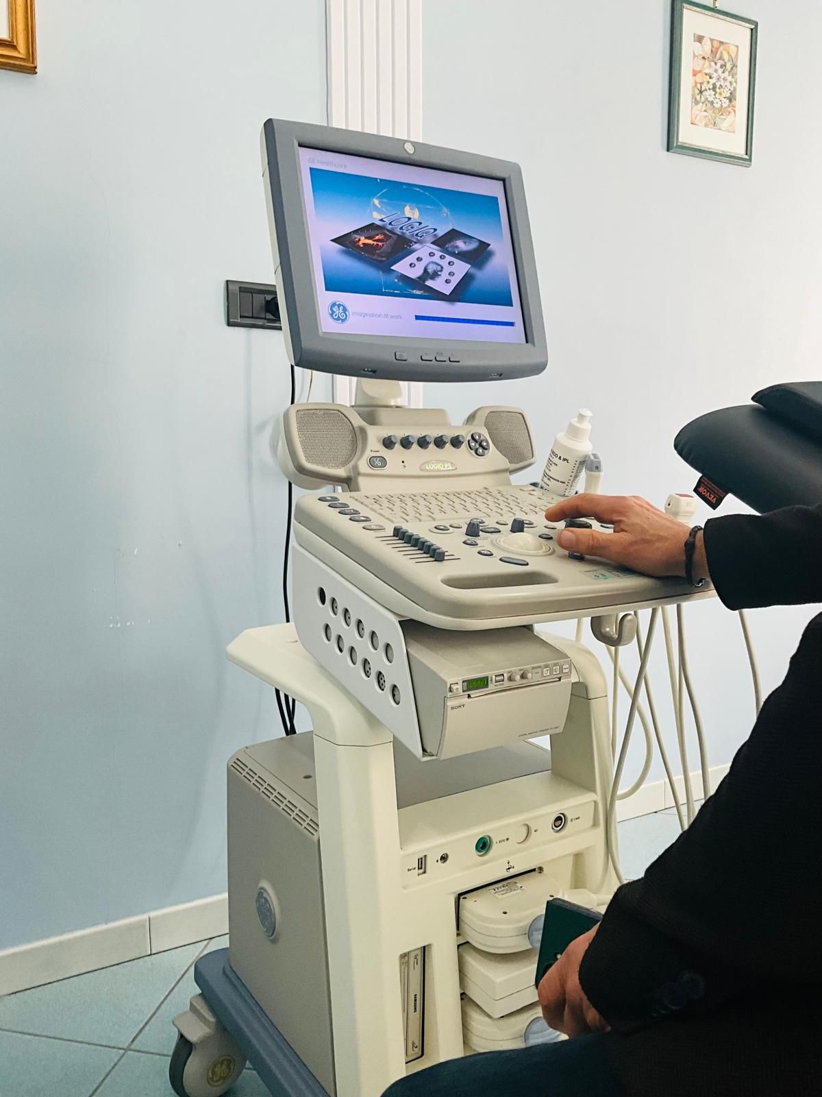 Ecografo accanto a un lettino bianco per visite in un ambiente medico.