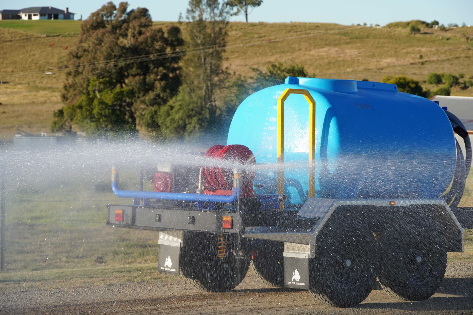 The 2000L poly water tank  of the water trailer in action while it is spraying water over a dirt road.