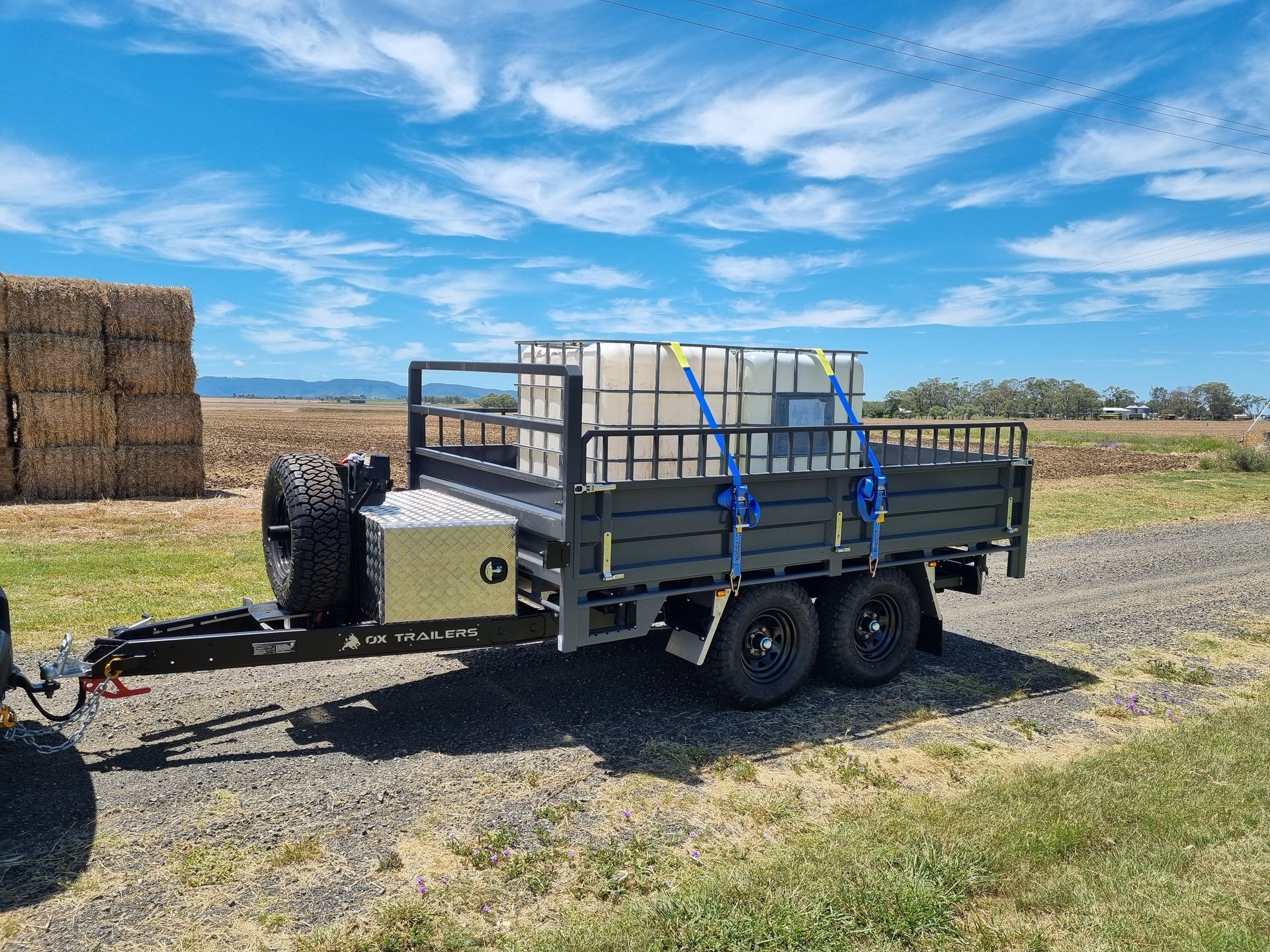 A 3-way tipper trailer hauling a poly oil tank on a gravel road with a large haystack in the background.