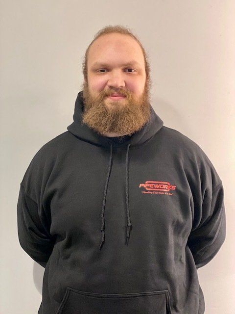 A man with a beard wearing a black hoodie, stands in front of a gray wall. He is smiling.