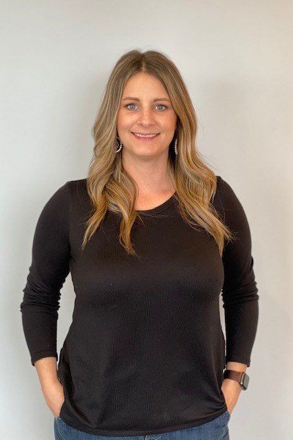 Woman with long blonde hair wearing a black long-sleeved shirt and blue jeans, standing in front of a white wall.