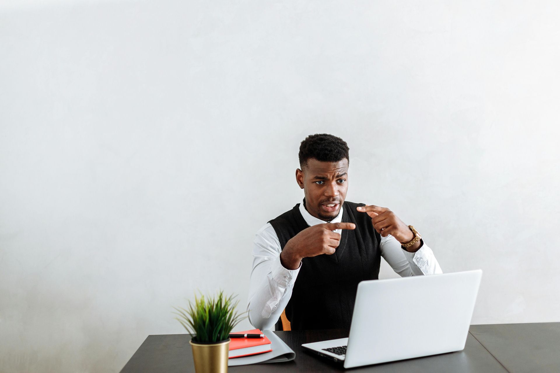 Man in white shirt and vest points angrily at laptop screen, seated at desk, with potted plant and books.