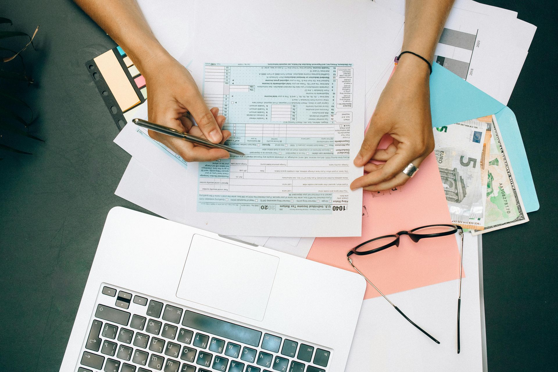 Hands filling out tax forms at a desk with a laptop, glasses, cash, and documents.
