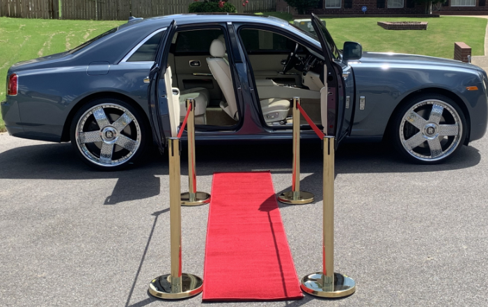 Dark blue luxury sedan with open door on a red carpet, framed by gold stanchions