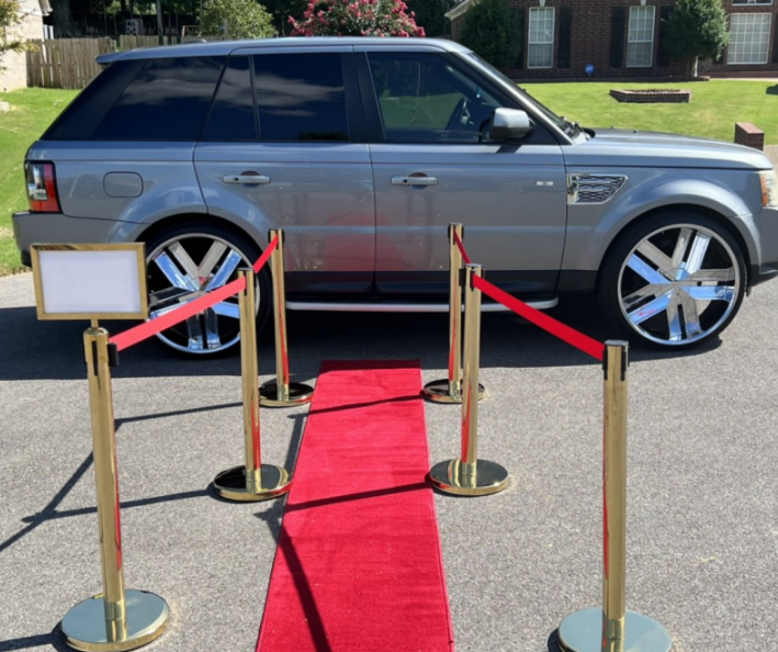 Silver SUV on display behind red carpet and gold stanchions outdoors.