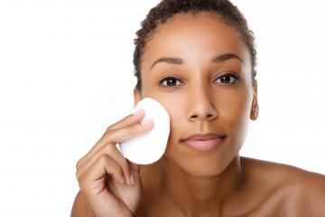 Person applying a white cotton pad to their cheek against a plain white background