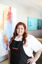Smiling person in a black apron and white shirt standing in an art studio with colorful abstract paintings.