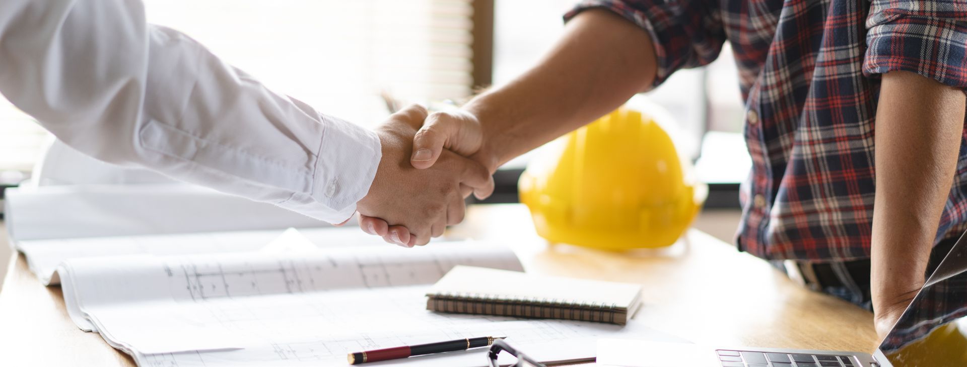 Two men are shaking hands over a table with a hard hat in the background.