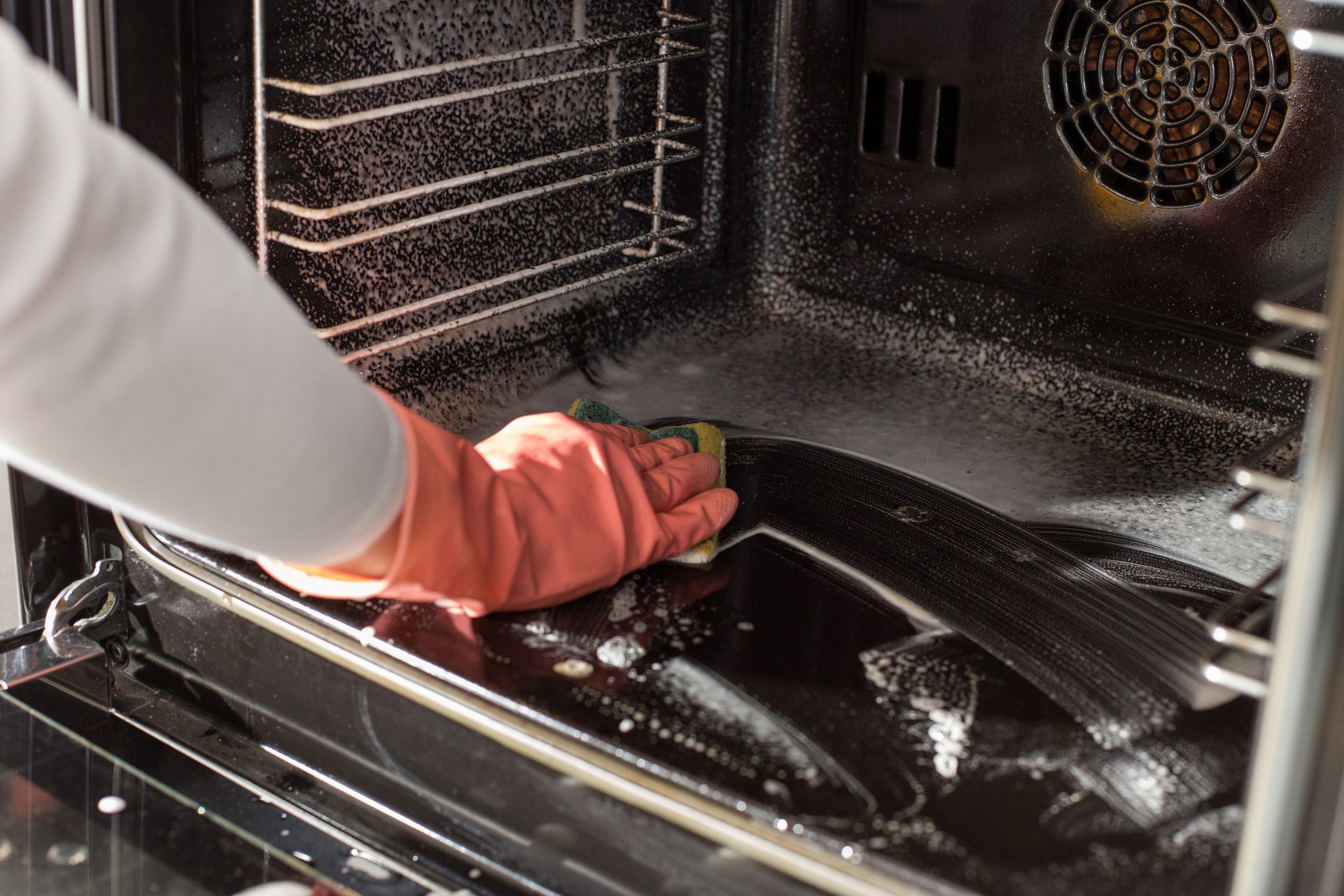 Person in orange gloves cleaning a messy oven interior with a sponge.