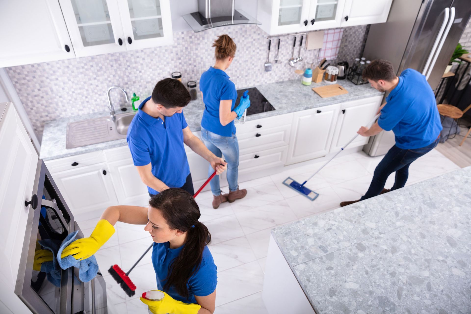 Four people wearing blue shirts cleaning a white kitchen.
