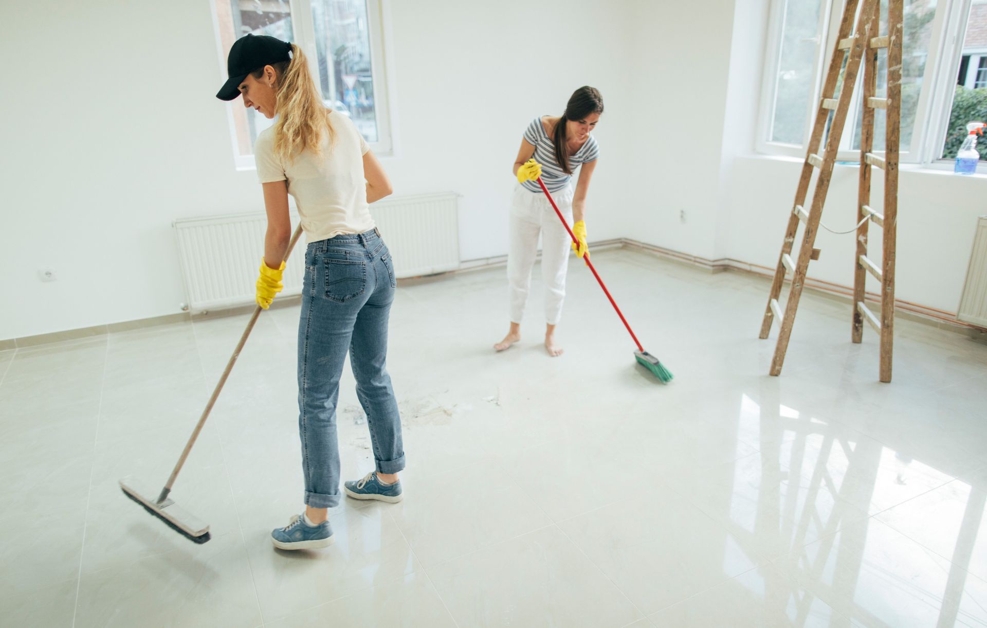 Two women cleaning a room with mops and brooms; white walls, windows, and ladder.