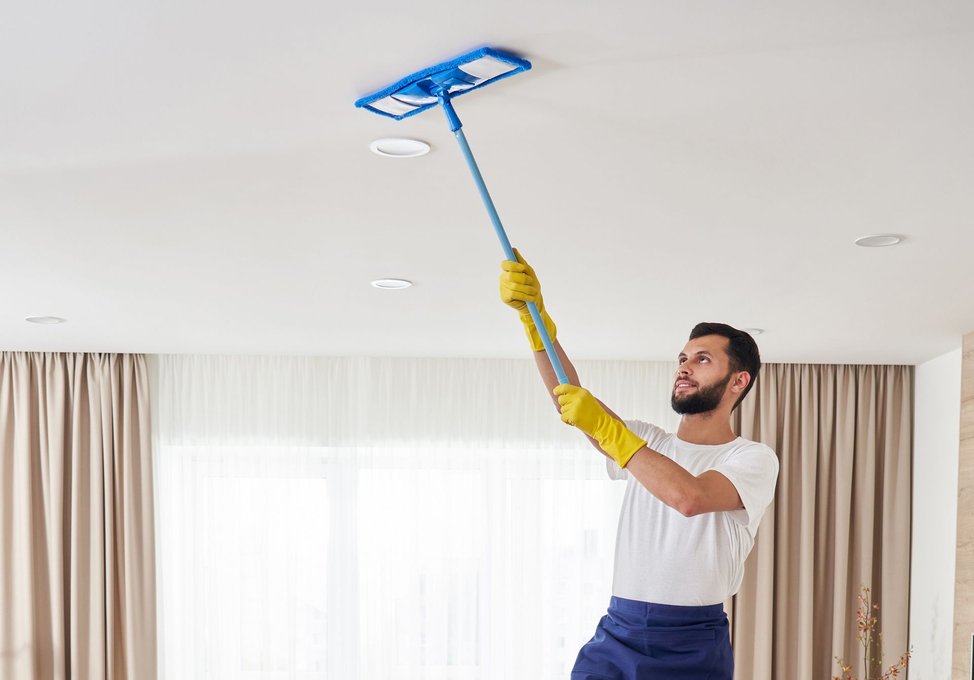 Man on a ladder, wearing gloves, cleans a ceiling with a blue mop.