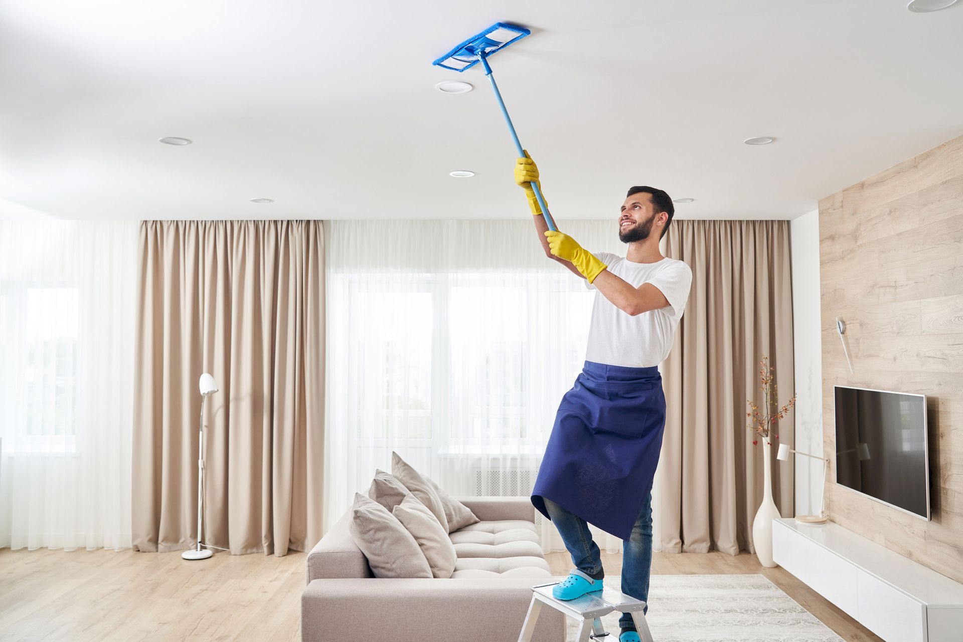 Man cleaning ceiling with a long-handled duster, standing on a ladder in a living room.
