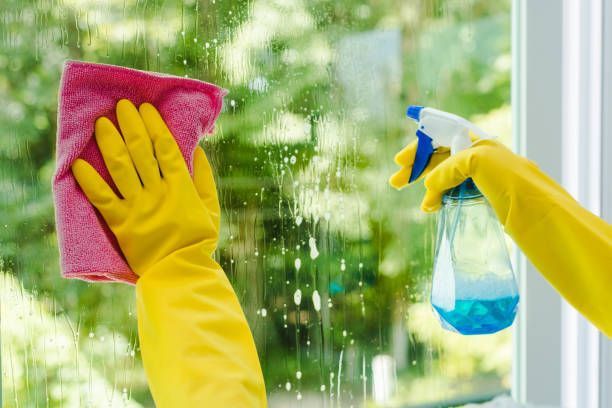 Person wearing yellow gloves sprays and wipes a window with a pink cloth.