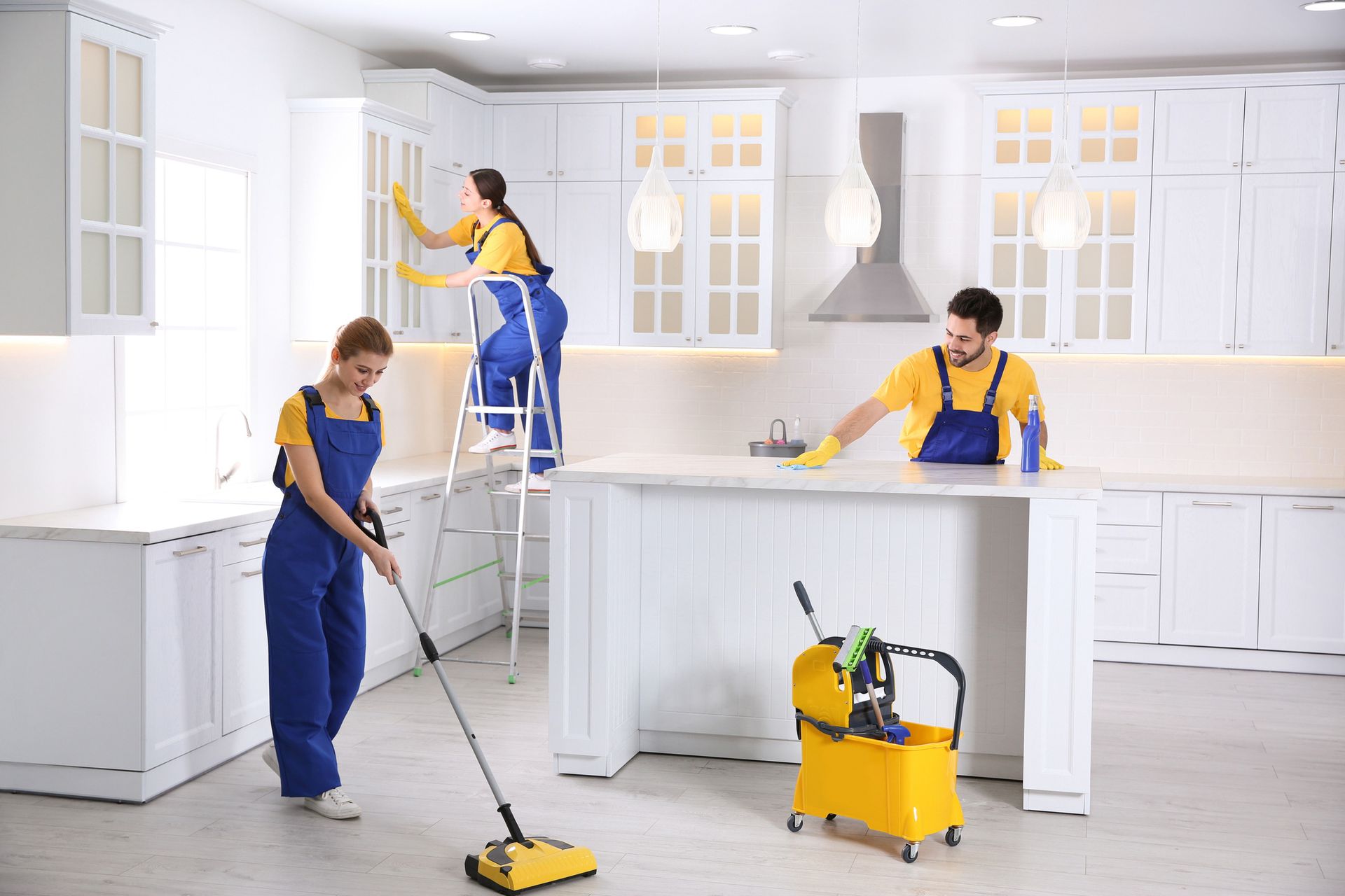 Three cleaners in a white kitchen, one on a ladder wiping cabinets, others cleaning surfaces and floor.