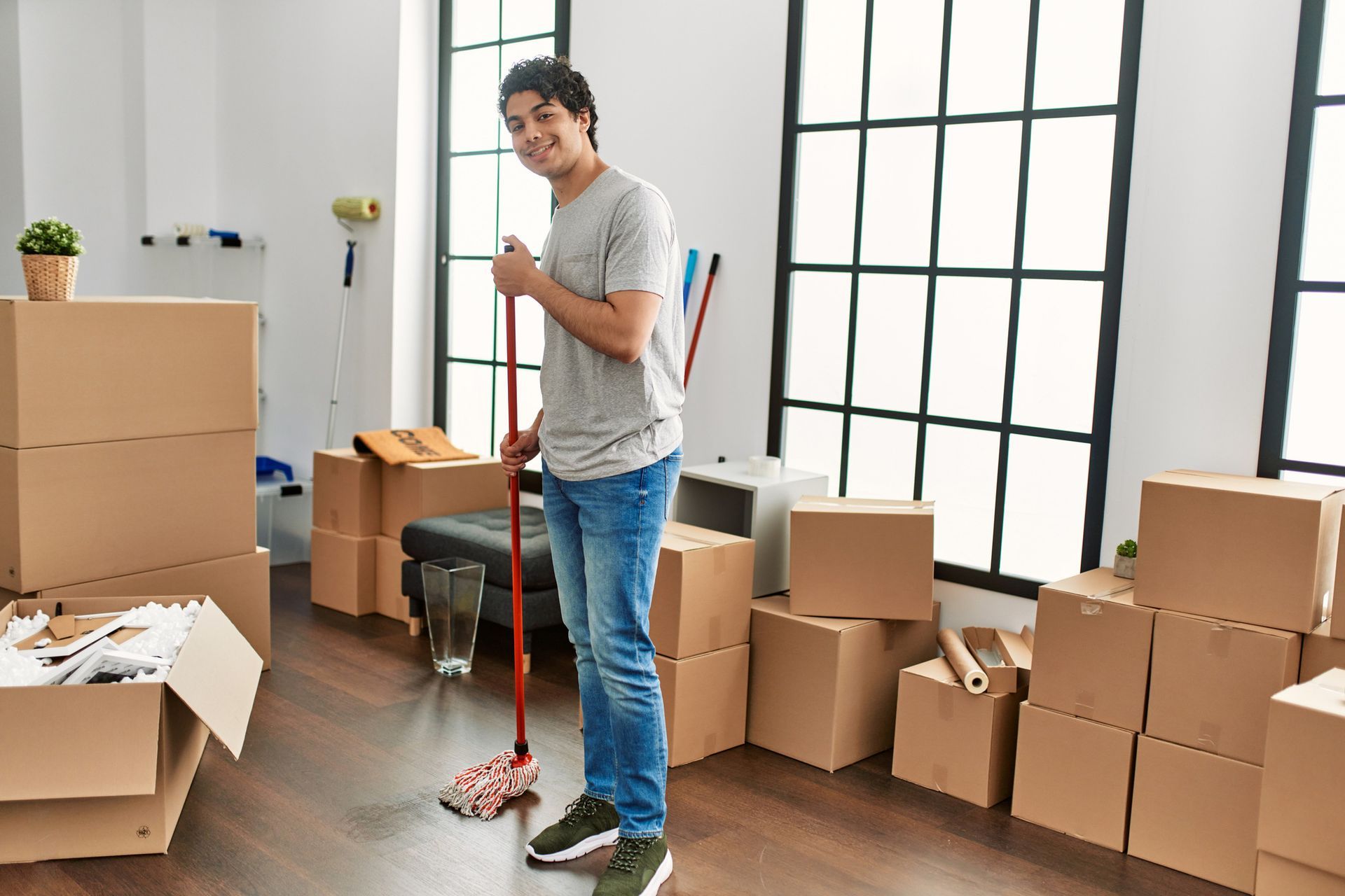 Man mopping floor in room with moving boxes, smiling at camera.