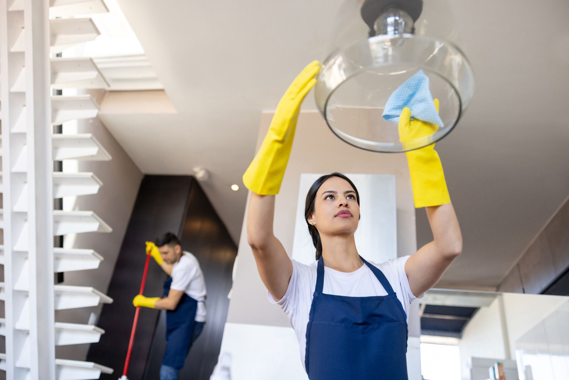 Woman in gloves cleans light fixture; another mops in background of modern home.