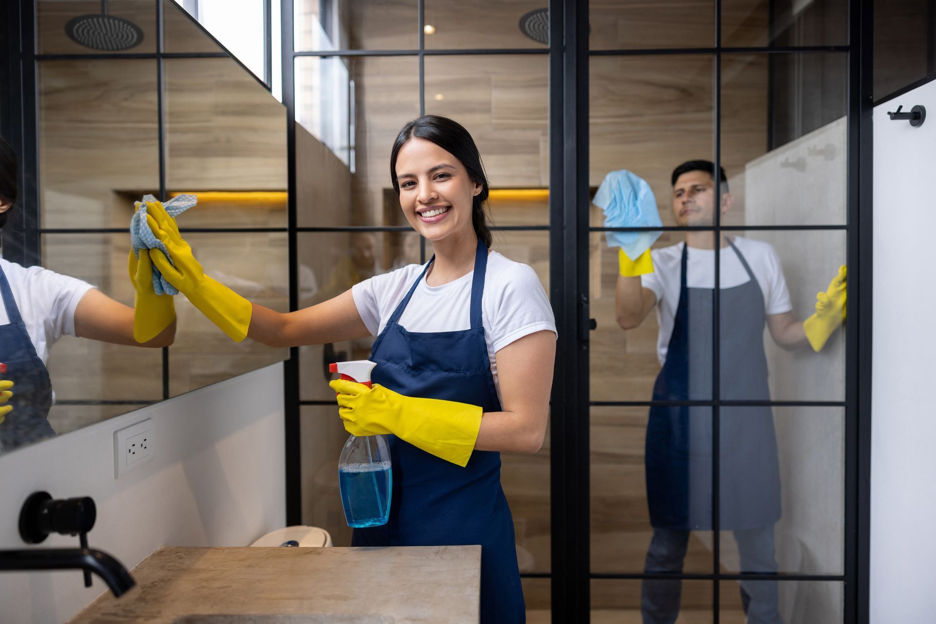 Woman and man wearing aprons and gloves cleaning a bathroom mirror.