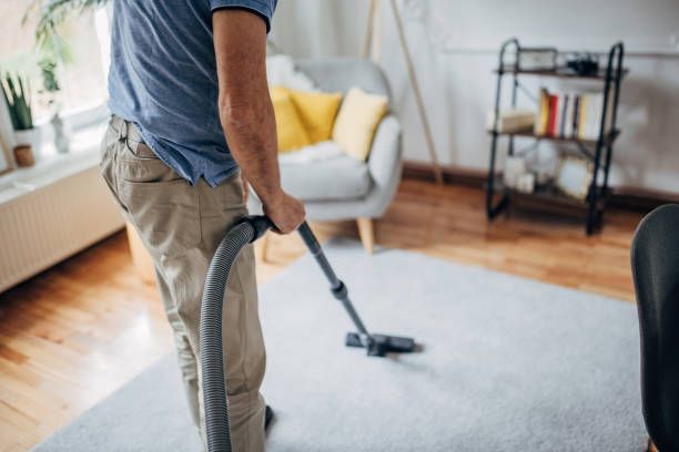 Person vacuuming a light-colored rug in a living room. The room has a chair, shelf, and wood flooring.