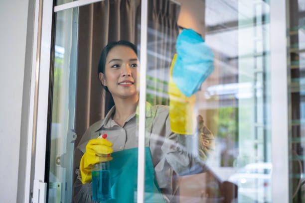 Woman cleaning a window, wearing yellow gloves and holding a spray bottle, smiling.